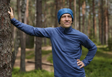 Load image into Gallery viewer, Man wearing a blue GOG quilted down beanie, leaning against a tree trunk in a sunlit pine forest with a hand resting on his hip.