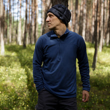 Load image into Gallery viewer, Man wearing a black GOG quilted down beanie, standing with a hand on a tree trunk in a sunlit pine forest.